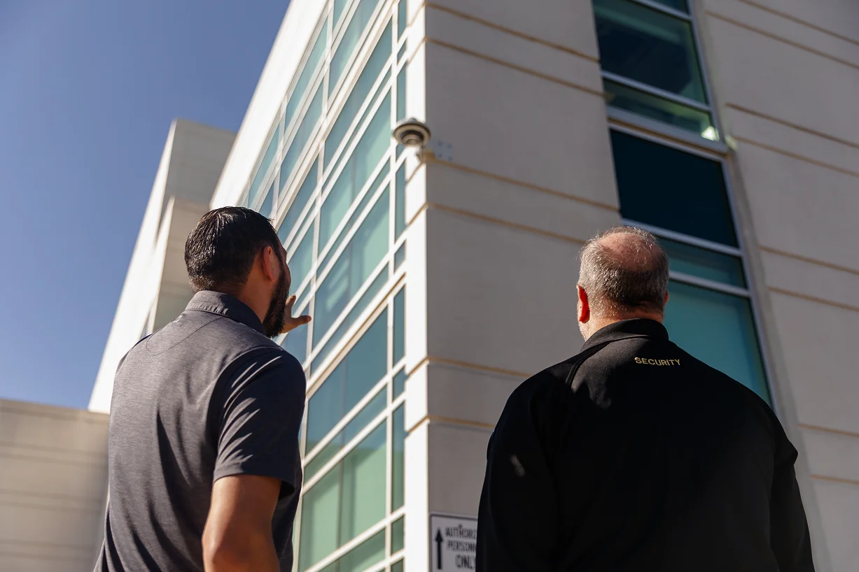 two men gesturing toward exterior camera outside of campus building